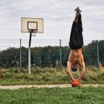 Homem faz parada de mãos equilibrando uma bola de basquete em uma quadra ao ar livre.