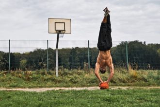 Homem faz parada de mãos equilibrando uma bola de basquete em uma quadra ao ar livre.