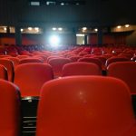 Sala de cinema vazia, com fileiras de poltronas vermelhas diante da tela iluminada.