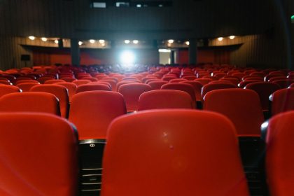 Sala de cinema vazia, com fileiras de poltronas vermelhas diante da tela iluminada.