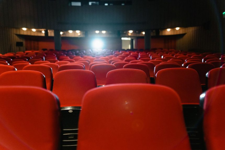 Sala de cinema vazia, com fileiras de poltronas vermelhas diante da tela iluminada.